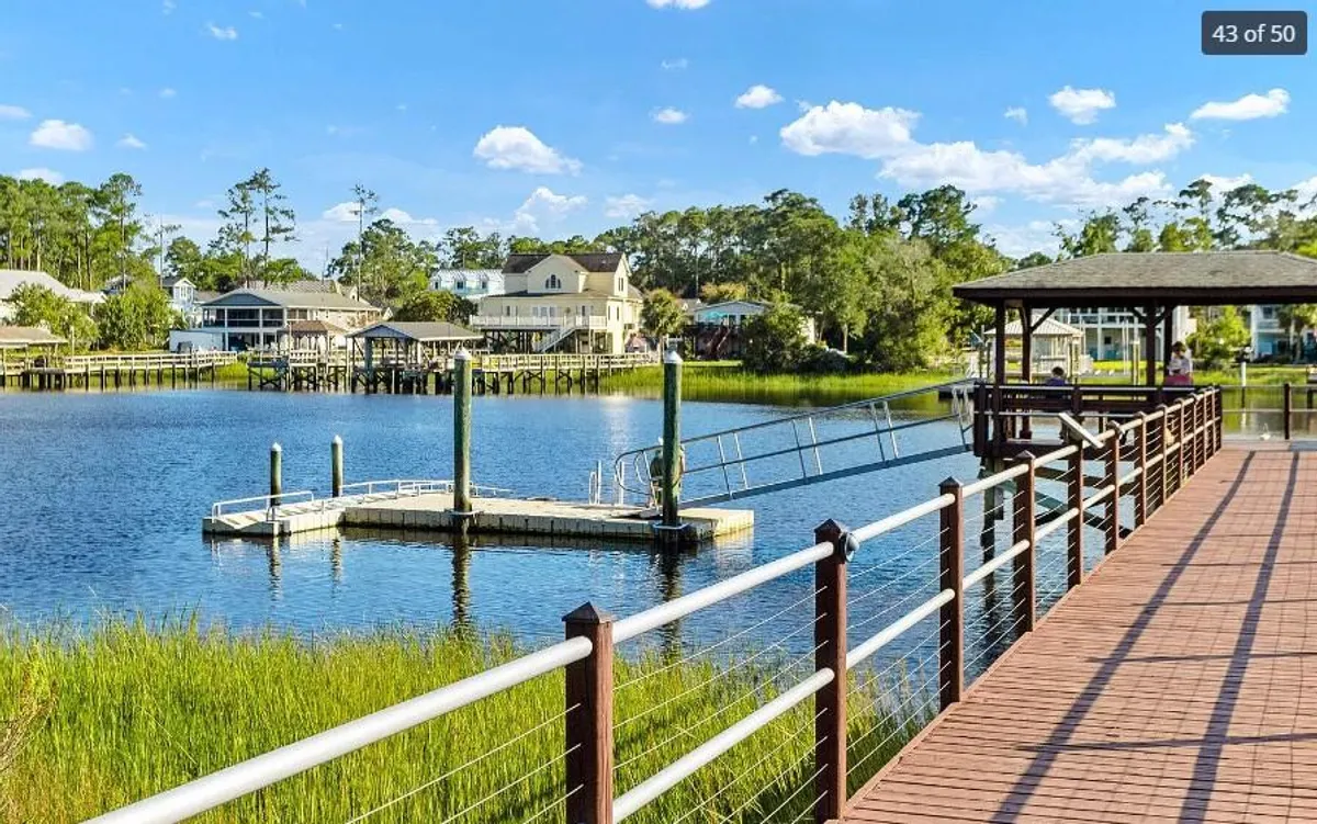 This image features a serene dock extending over calm water, with houses and a boardwalk visible in the background. The setting evokes a peaceful waterfront atmosphere, perfect for leisurely walks, boating, or simply soaking in the views at sunset.