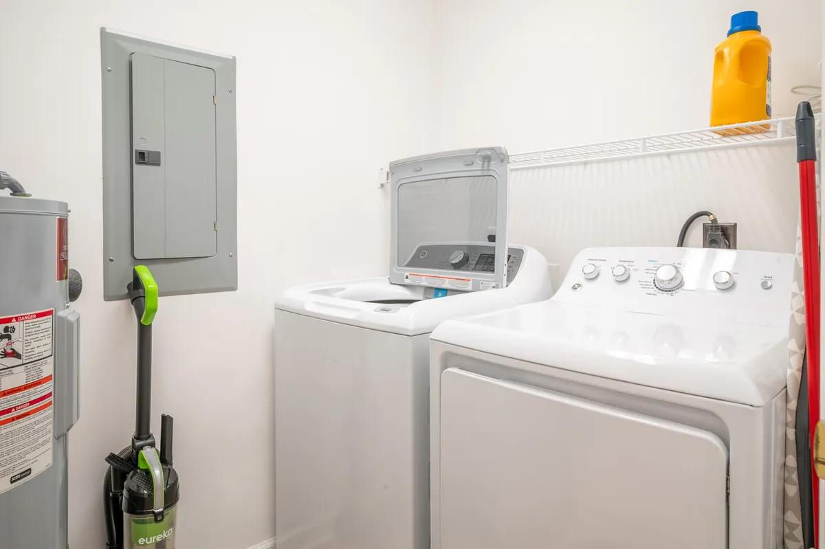 This practical laundry area includes a washing machine, dryer, cleaning supplies, and an electrical panel, all neatly arranged. The setup conveys functional convenience and is ideal for guests planning extended stays who appreciate having in-unit laundry facilities.