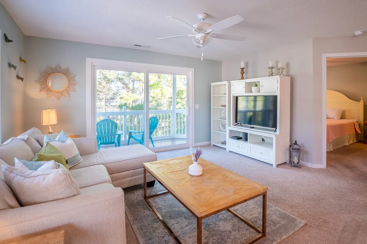 A bright and comfortable living space with a sofa, coffee table, Smart TV, and a sliding glass door leading to an outdoor balcony with seating. The room is designed for both relaxation and entertainment, with a clear view to the outdoors.