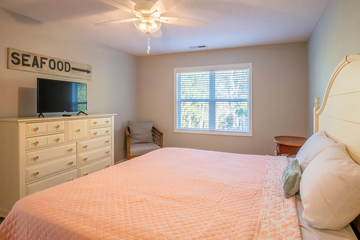 This image features a tranquil bedroom with a plush bed dressed in a pink quilt, a dresser topped with a television, and a window letting in natural light. The room is styled simply, evoking comfort and serenity, making it an inviting retreat for rest or catching up on favorite shows. The television and bed are the main focal points, directly supporting the narrative of relaxation and entertainment within the private suite.
