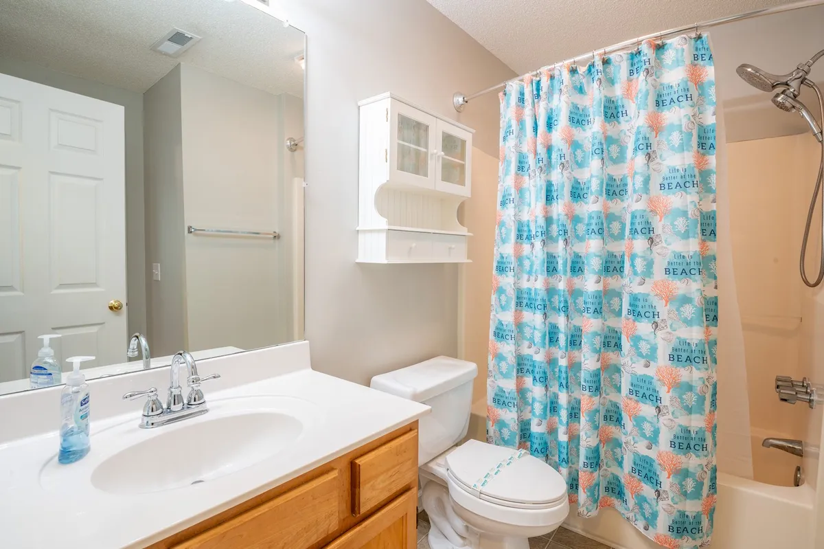 A bright, welcoming bathroom scene featuring a vanity with sink, a mirrored cabinet, a beach-themed shower curtain concealing a step-in shower, and a toilet. The decor is light and casual, with practical storage elements visible, capturing the section’s emphasis on ensuite convenience and practical amenities.