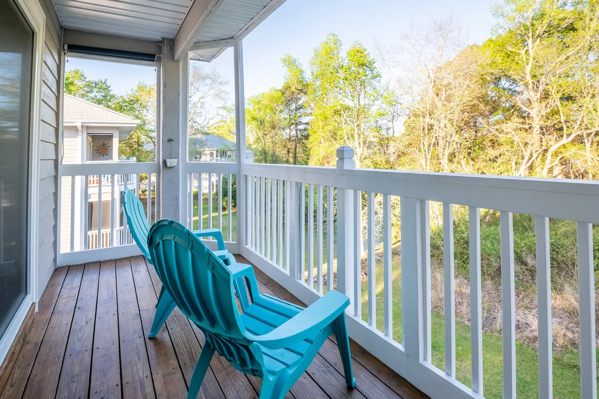 A wooden deck with blue Adirondack chairs faces a backdrop of lush green trees, inviting guests to relax, read, or enjoy the peaceful sounds and breezes from the coast. The scene captures the essence of tranquil outdoor leisure moments.